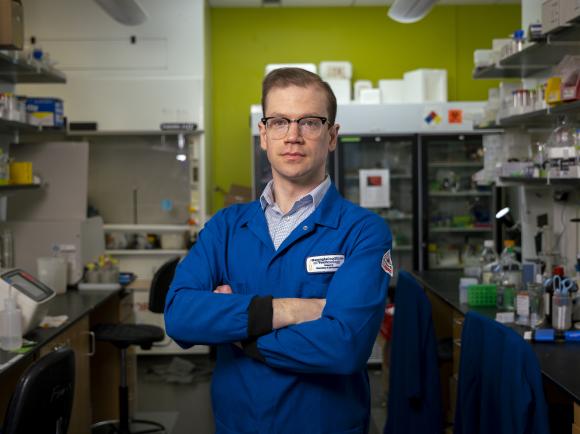 A person wearing a blue lab coat stands with arms crossed in a laboratory filled with shelves of scientific equipment, supplies, and a refrigerator unit in the background.