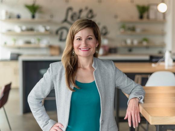 Alison Sizer in a blazer standing in a modern workspace with wooden tables, open shelving, and natural light.