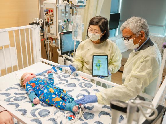 Two adults wearing protective gowns and gloves stand beside a hospital crib, using a tablet device while examining an infant lying on the mattress as medical equipment and monitors surround the crib.