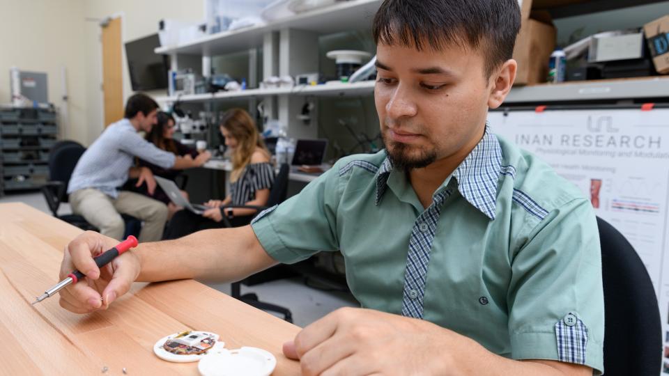 A man with tan skin and dark hair, wearing a mint-green shirt, is seated at a table and looking at the CardioTag device.